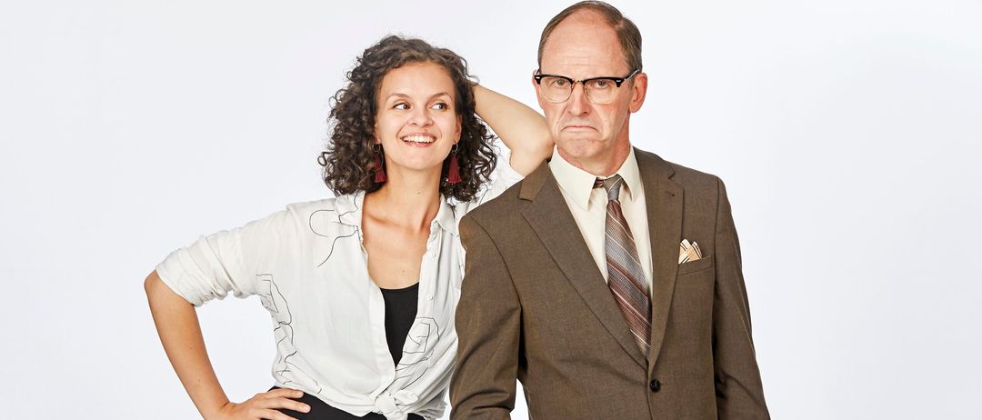 Eine Frau mit lockigem Haar lächelt und posiert mit der Hand am Kopf. Neben ihr steht ein älterer Mann in einem Anzug, der eine Brille und eine Krawatte trägt.