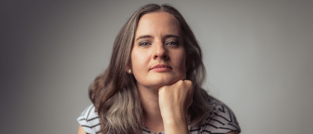 Eine Frau mit grauen Haaren schaut in die Kamera. Sie trägt ein gestreiftes Hemd und hat die rechte Hand im Kinn. Sie hat ein Armband am linken Handgelenk. Sie ist wahrscheinlich in einem Studio.