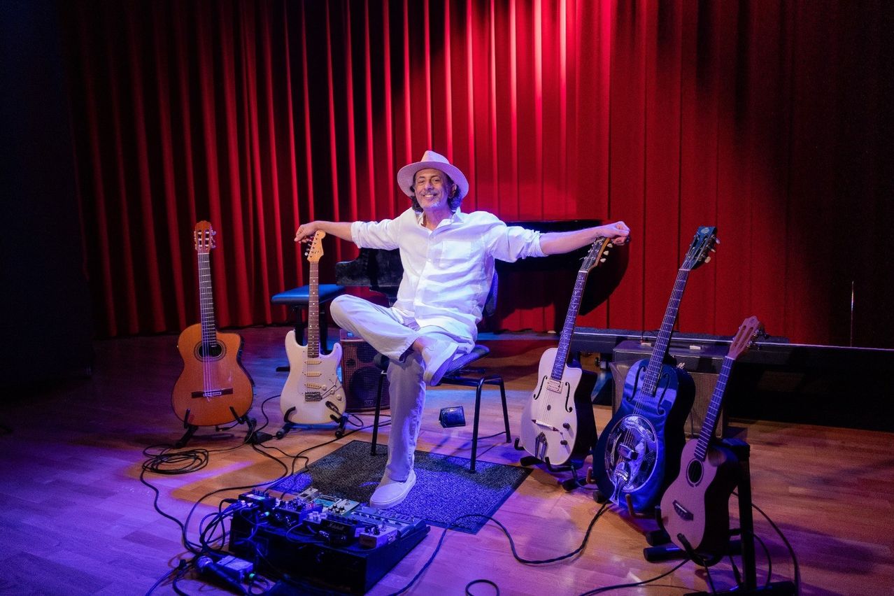 Ein Mann sitzt auf einem Hocker vor mehreren Gitarren und einem Klavier, trägt einen Hut und ein Lächeln. Zu den Gitarren gehören eine klassische, eine Stahlsaiten-Akustik und eine Resonator. Im Hintergrund hängen rote Vorhänge.