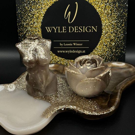 Zwei dekorative Objekte befinden sich auf einem goldfarbenen Tablett. Die Objekte sind eine Büste und eine Rose. Die Büste befindet sich links und die Rose rechts. Die Objekte sind mit Goldglitter bedeckt. Der Hintergrund ist ein schwarzes Kästchen mit Goldglitter.