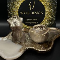 Zwei dekorative Objekte befinden sich auf einem goldfarbenen Tablett. Die Objekte sind eine Büste und eine Rose. Die Büste befindet sich links und die Rose rechts. Die Objekte sind mit Goldglitter bedeckt. Der Hintergrund ist ein schwarzes Kästchen mit Goldglitter.