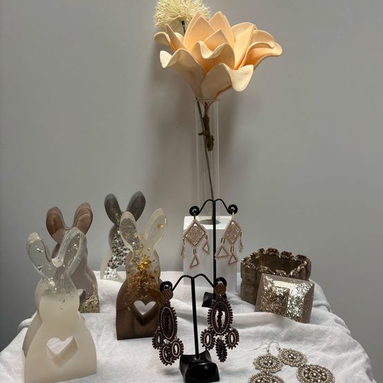 Ein mit einem weißen Tuch bedeckter Tisch, auf dem Schmuck ausgestellt ist, darunter Hasenfiguren, Ohrringe und Armbänder. Hinter dem Schmuck steht ein großes beigefarbenes Blumenarrangement in einer Vase.