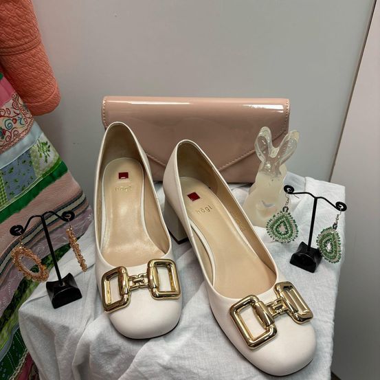 Ein Paar weiße flache Schuhe mit goldenen Schnallen und einer rosa Handtasche sind auf einem weißen Tuch ausgestellt.
