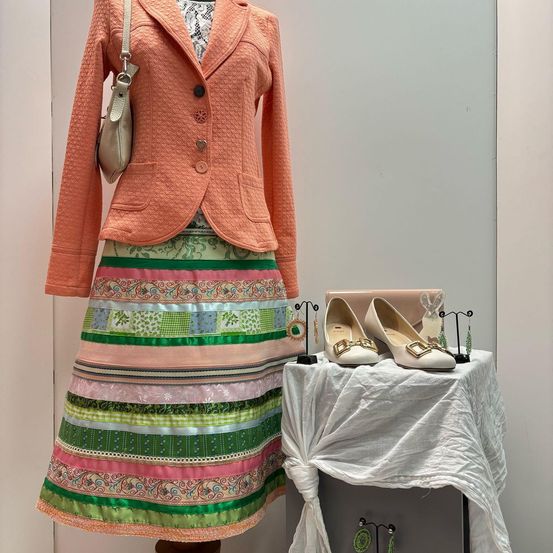 Eine Modeschau mit einer pfirsichtfarbenen Jacke, einem mehrfarbigen Rock und einem Paar weißer Schuhe auf einem mit weißem Stoff bedeckten Tisch.