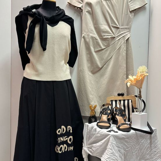 Ein schwarz-weißer Rock mit goldenen Buchstaben und ein weißes Oberteil mit schwarzem Schal. Ein beiges Kleid und schwarze Sandalen stehen auf einem Tisch mit einer Handtasche und einer Blume.