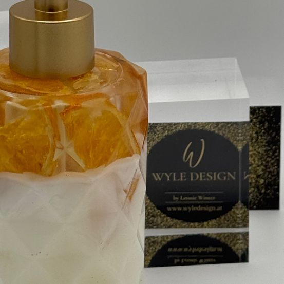 Ein goldener Seifenspender mit Marmordesign steht auf einer weißen Oberfläche, mit einer durchsichtigen Glasbox und einem schwarzen Etikett mit dem Text WYLE DESIGN.