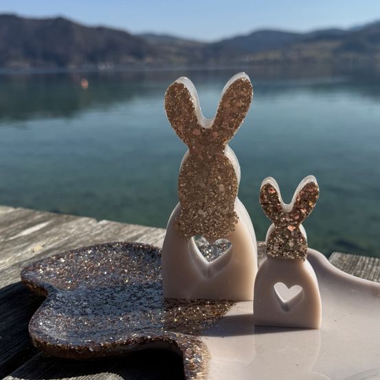 Zwei dekorative Hasenfiguren mit Glitzer auf einer Holzoberfläche, in der Nähe eines ruhigen Sees mit Bergen im Hintergrund.