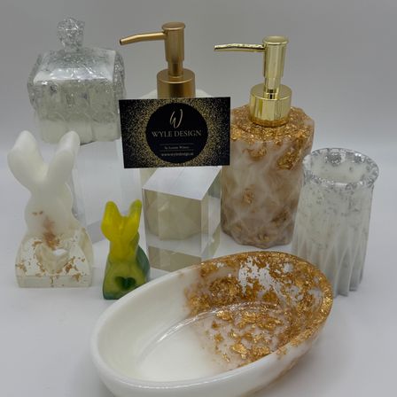 Verschiedene dekorative Objekte und Badezimmerzubehör auf einer weißen Oberfläche, einschließlich eines goldenen Seifenspenders, einer Schüssel, eines Bechers, einer Box und einer Hasenfigur.