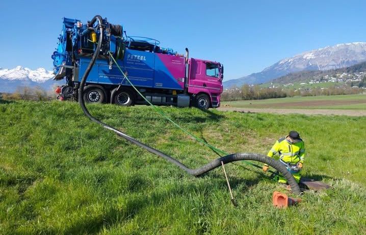 Ein Mann benutzt einen großen Schlauch, der mit einem blauen und rosa Truck verbunden ist, um Wasser in den Boden in einem Grasfeld zu pumpen.