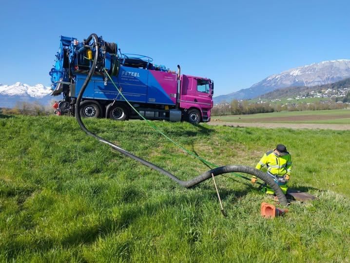 Ein Mann benutzt einen großen Schlauch, der mit einem blauen und rosa Truck verbunden ist, um Wasser in den Boden in einem Grasfeld zu pumpen.