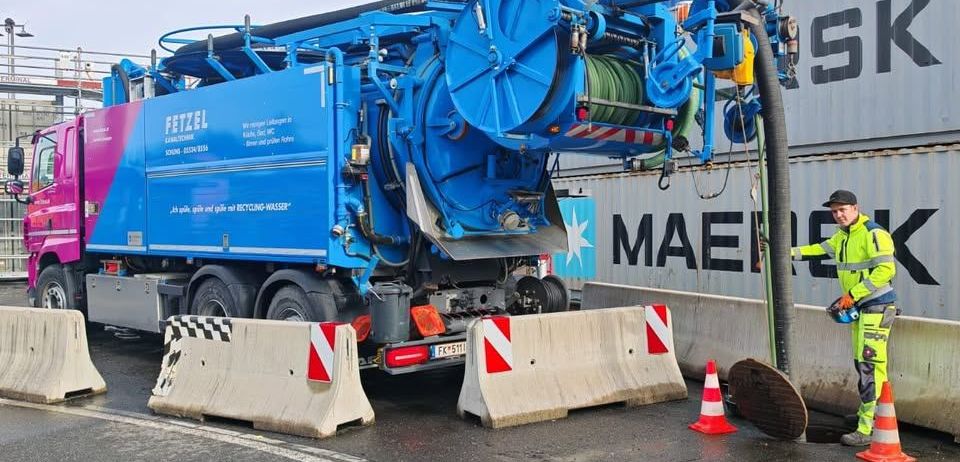 Ein blauer Recycling-LKW steht am Straßenrand, bedient von einem Mann in einem gelben Overall. Er hat einen großen, zylindrischen Tank auf dem Rücken.