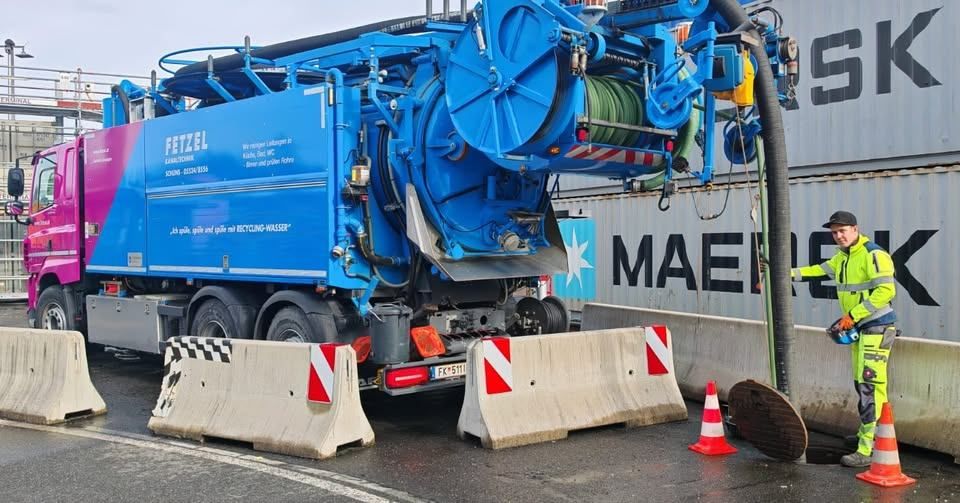 Ein blauer Recycling-LKW steht am Straßenrand, bedient von einem Mann in einem gelben Overall. Er hat einen großen, zylindrischen Tank auf dem Rücken.