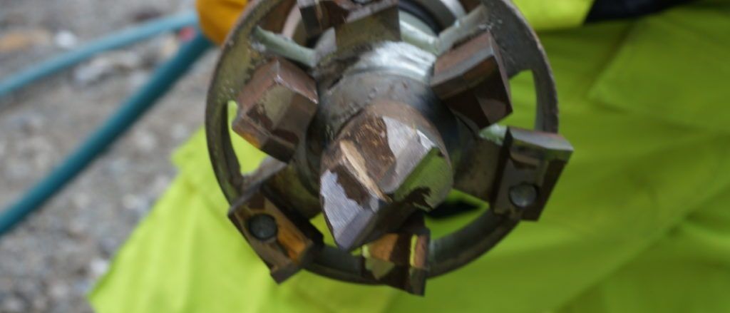 Eine Nahaufnahme eines metallischen Teils einer Bohrmaschine, das von einem Arbeiter in einer neongrünen Sicherheitsjacke mit gelben Handschuhen gehalten wird.