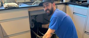 Ein Mann in blauem Hemd und gelber Hose kniet unter einem Waschbecken, hält eine Rohrleitung fest und neben ihm steht ein Gerät auf dem Boden.