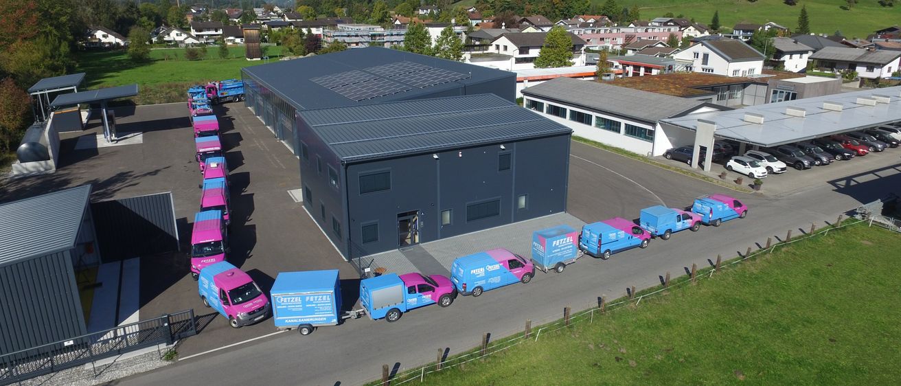 Ein Luftbild einer Anlage mit mehreren vor ihr geparkten Trucks, einige mit dem Wort 'Fetzer' darauf. Das Gebiet ist von einem Zaun umgeben und es gibt Häuser und einen Berg in der Ferne.