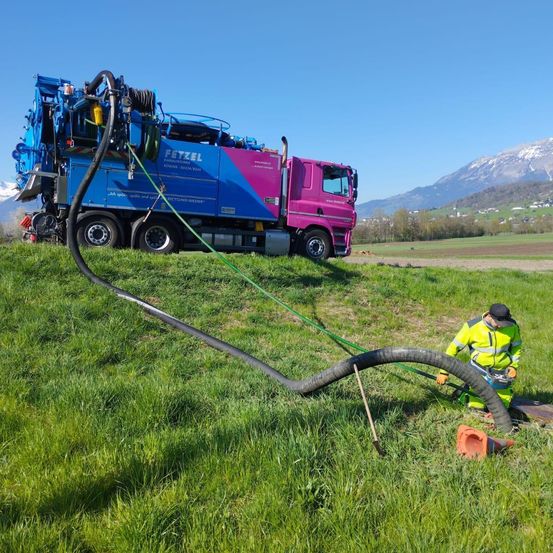 Ein Arbeiter bedient einen großen blauen und pinken Lastwagen mit angeschlossener Schläuche, auf einem Grasfeld geparkt. Im Hintergrund sind Berge zu sehen.
