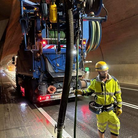 Ein Arbeiter in einer reflektierenden gelben Weste bedient einen großen Vakuumsauger in einem Tunnel. Der Lkw hat mehrere Schläuche und ist mit einer großen Rohrleitung verbunden.