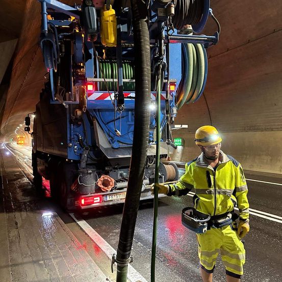 Ein Arbeiter in einer reflektierenden gelben Weste bedient einen großen Vakuumsauger in einem Tunnel. Der Lkw hat mehrere Schläuche und ist mit einer großen Rohrleitung verbunden.