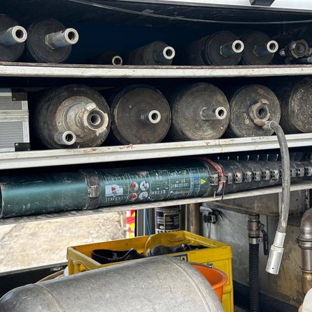 Ein Blick in einen Lkw mit mehreren Metallzylindern, einige mit angebauten Rohren. Ein großer grüner Zylinder mit Etiketten steht auf einem unteren Regal. Gelbe Aufbewahrungsboxen und ein roter Eimer sind im Vordergrund.