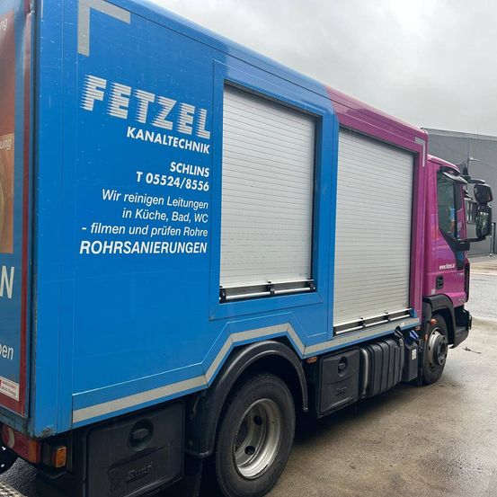 Ein blauer und rosafarbener LKW mit "FETZEL KANALTECHNIK" und "SCHLINS T 05524/8556" darauf, geparkt auf der Straße.