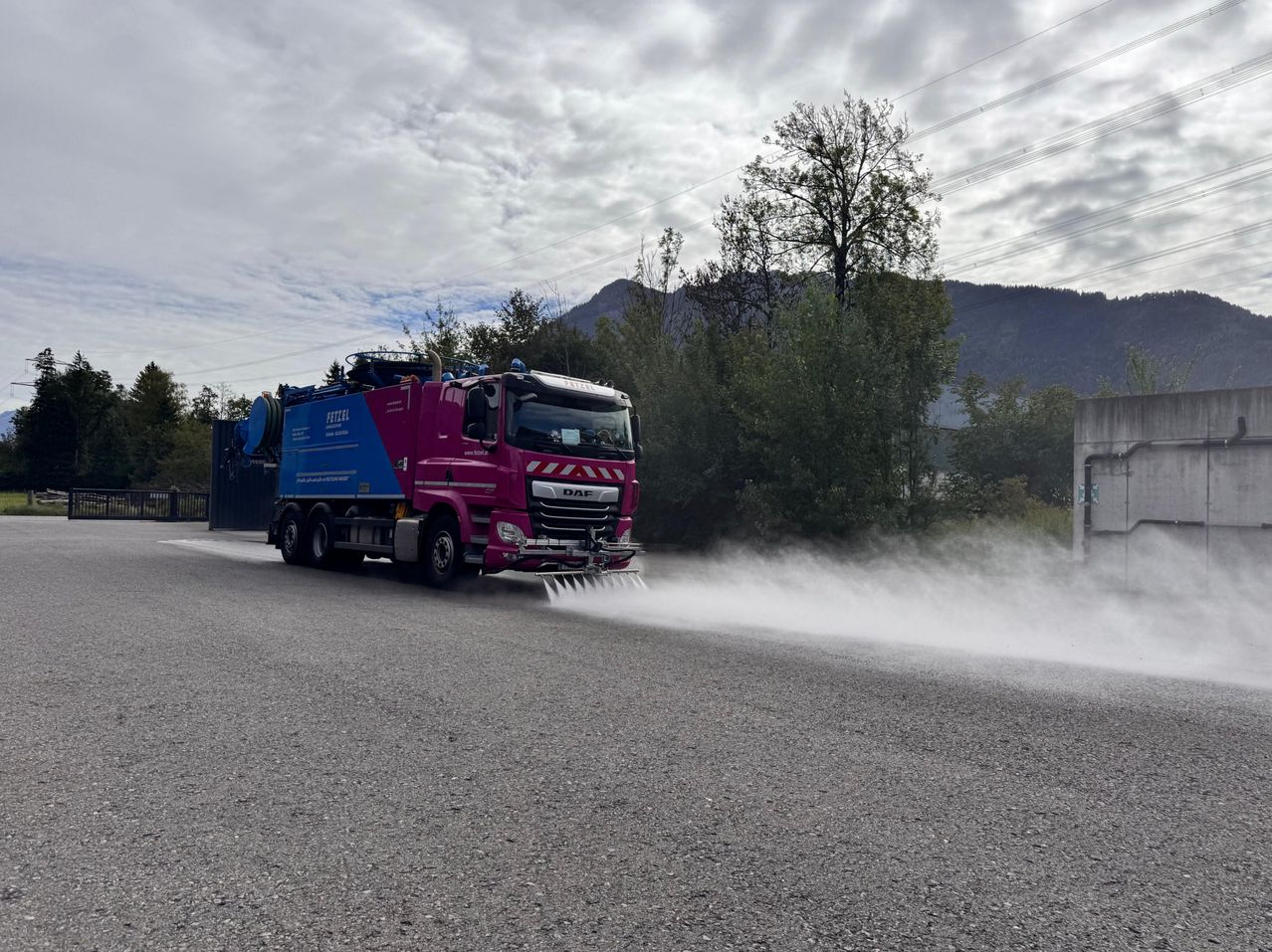 Ein lila und blauer DAF-Lkw sprüht Wasser auf die Straße. Er befindet sich auf einer Straße mit Bergen und Bäumen im Hintergrund.