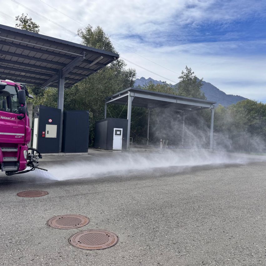Ein rosa Lkw sprüht Wasser auf die Straße vor einem Unterstand mit Solardach. Im Hintergrund befinden sich Kanaldeckel und Berge.