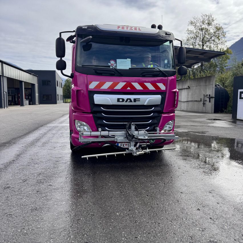 Ein rosafarbener DAF-Lkw mit montiertem Gerät steht auf einer nassen Straße, mit Gebäuden und Bäumen im Hintergrund.
