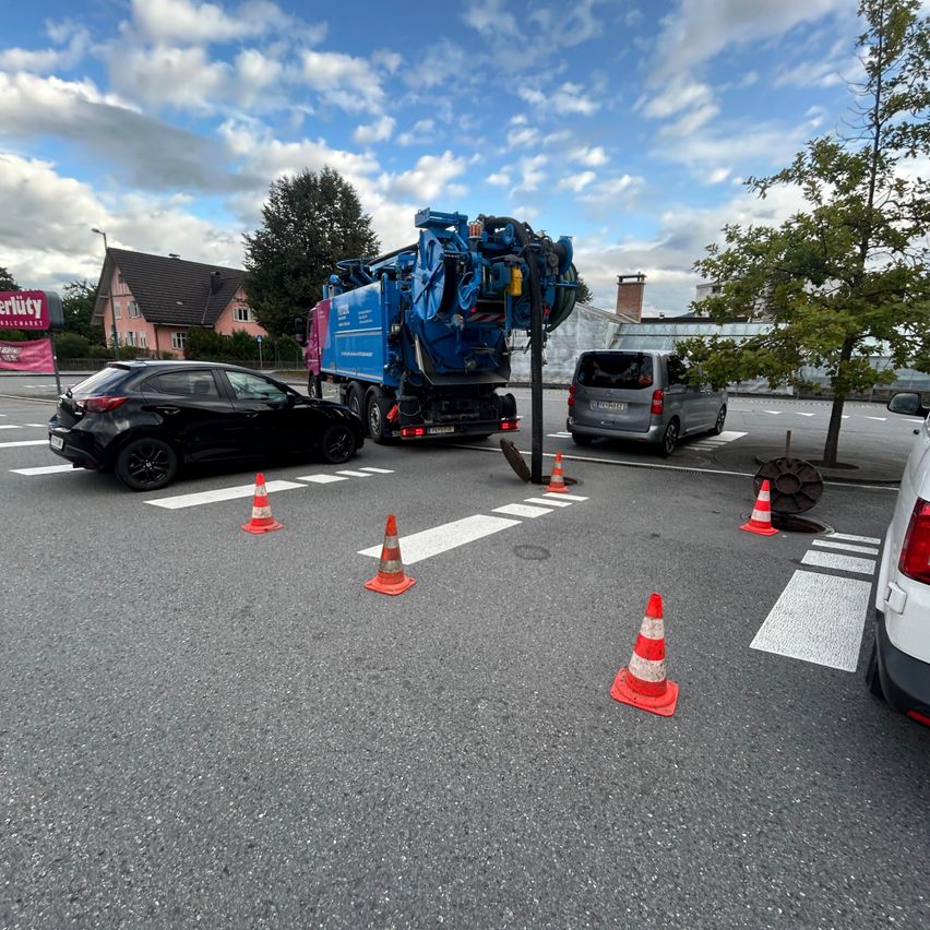 Ein blauer Lastwagen mit einer Reinigungsvorrichtung steht auf einer Straße. Ein Auto und ein Van sind in der Nähe. Es gibt orangefarbene Kegel und Zebrastreifen auf der Straße.