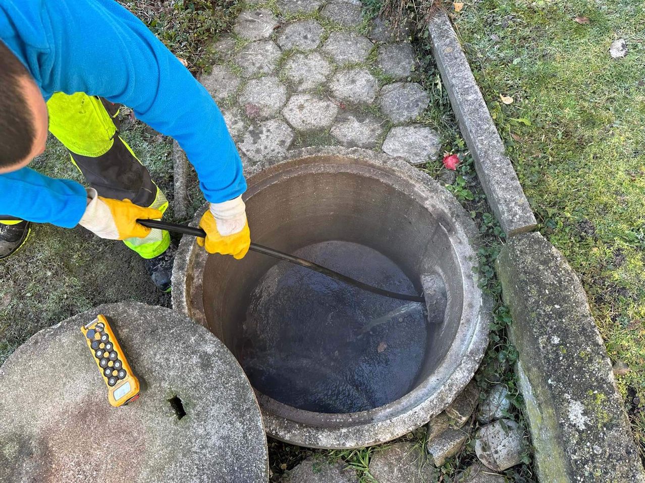 Ein Arbeiter in einem blauen Shirt und gelben Handschuhen benutzt ein langes Werkzeug, um einen großen, offenen Kanaldeckel zu überprüfen. In der Nähe befindet sich ein kleines gelbes Gerät mit Tasten.
