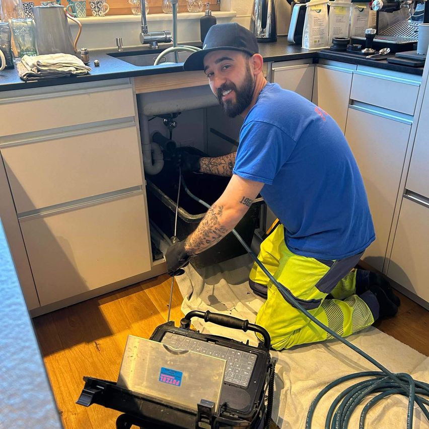 Ein Mann repariert einen Abfluss in einer Küche mit einem Werkzeug. Er trägt ein blaues Hemd und gelbe Hosen.