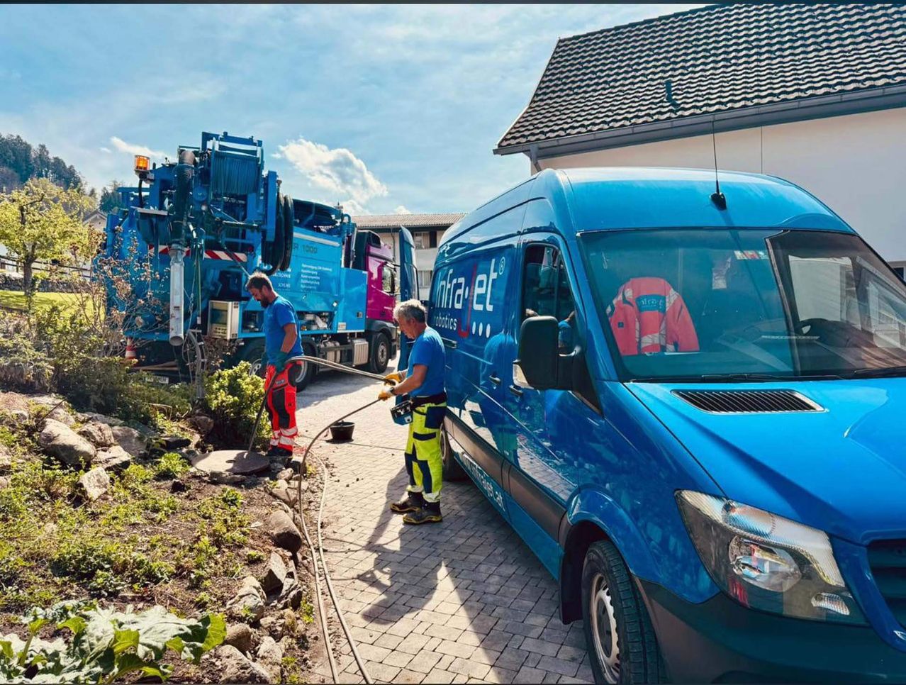 Zwei Männer in Schutzkleidung reinigen die Einfahrt mit einem Schlauch. Ein blauer Lastwagen mit dem Wort 'infra' ist in der Nähe geparkt.