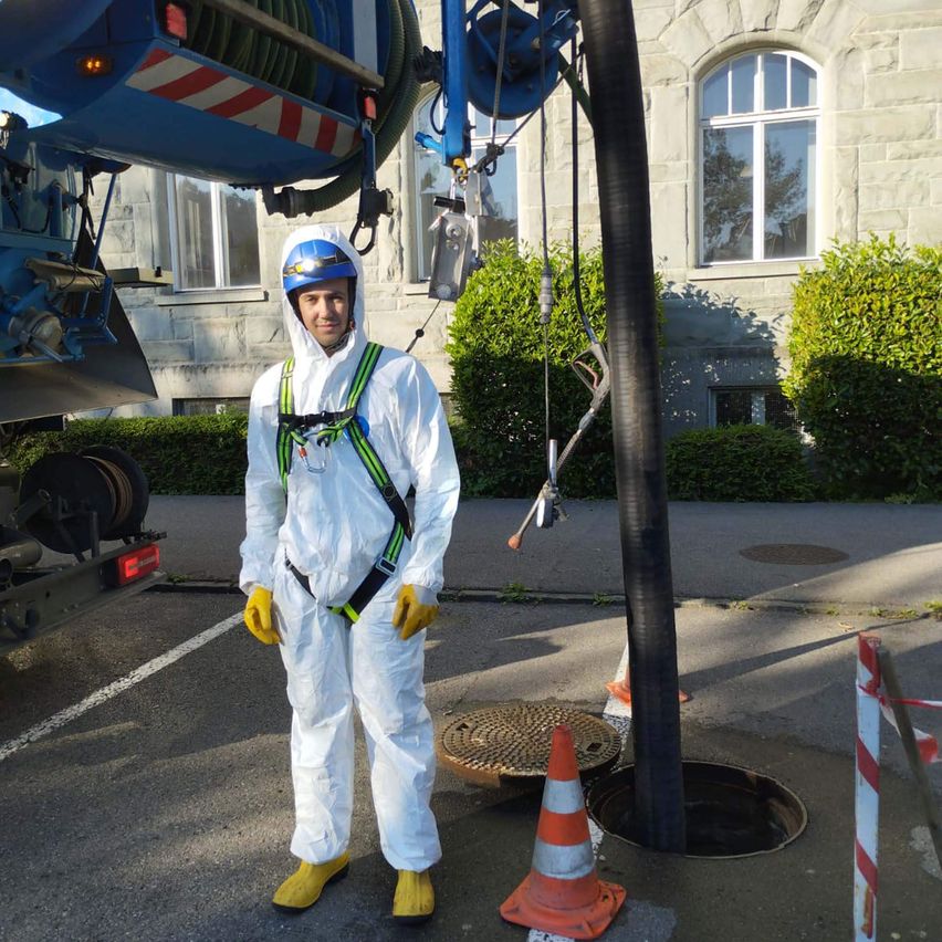 Ein Mann in einem weißen Schutzanzug und Helm steht in der Nähe eines Abwasserrostes, mit einer großen blauen Maschine dahinter und einem orangefarbenen Verkehrskegel in der Nähe.
