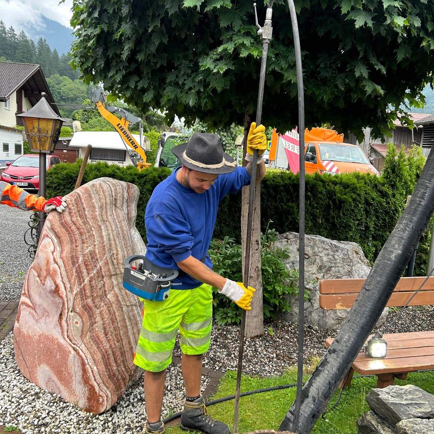 Ein Mann in einem blauen Shirt, gelben Handschuhen und grüner Shorts hält einen Pfahl, möglicherweise bei der Arbeit an einem Baum in einem Park. In der Nähe gibt es einen großen Felsen, eine Bank und ein Baufahrzeug.
