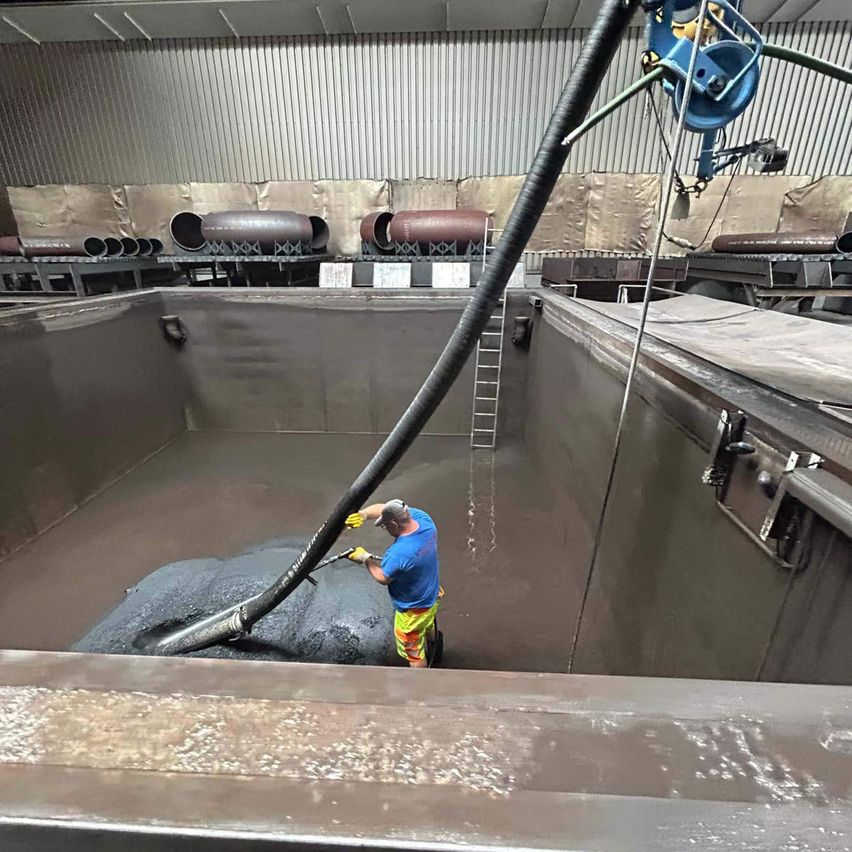 Ein Mann in einem blauen Shirt und gelber Hose reinigt Kies in einer großen Betongrube mit einem Schlauch. Die Grube befindet sich in einer Lagerhalle mit Metallwänden und Rohren.