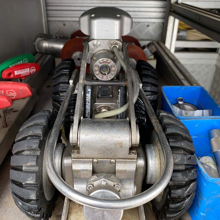 A metal vehicle with large black tires is displayed in a garage. Several tools are on the left side of the vehicle. Blue bins and shelves are on the right side.