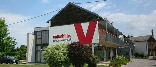 Ein modernes Gebäude mit einem roten und weißen Schild, auf dem 'volkshiffe. SENIORENZENTRUM' steht. Es hat eine Einfahrt und ein schwarzes Auto davor. Das Gebäude ist von Sträuchern und Pflanzen umgeben.