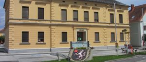Ein gelbes Gebäude mit mehreren Fenstern und einem Schild, das Rathaus steht. Ein kleiner Garten mit Blumen und einem dekorativen Emblem steht vor dem Gebäude.