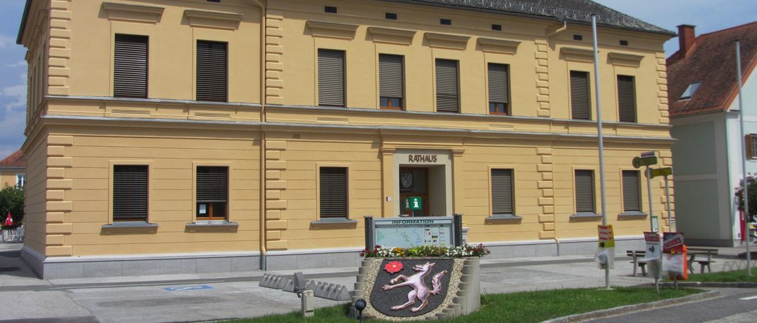Ein gelbes Gebäude mit mehreren Fenstern und einem Schild, das Rathaus steht. Ein kleiner Garten mit Blumen und einem dekorativen Emblem steht vor dem Gebäude.