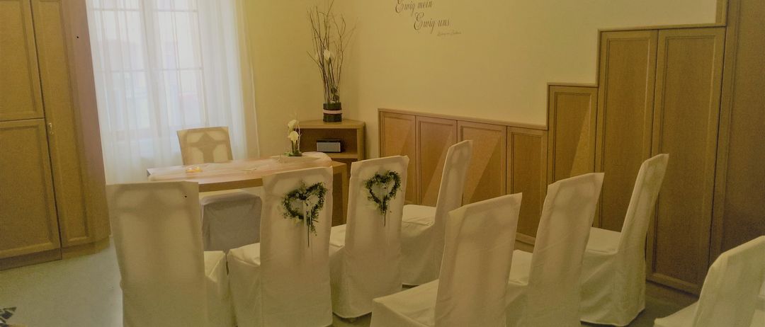 Ein Raum mit weißen Stühlen, einem hölzernen Schreibtisch und einer Wand mit Blumenarrangement und Text. Die Stühle sind in einer Reihe angeordnet, wobei einige dekorative Kränze haben.