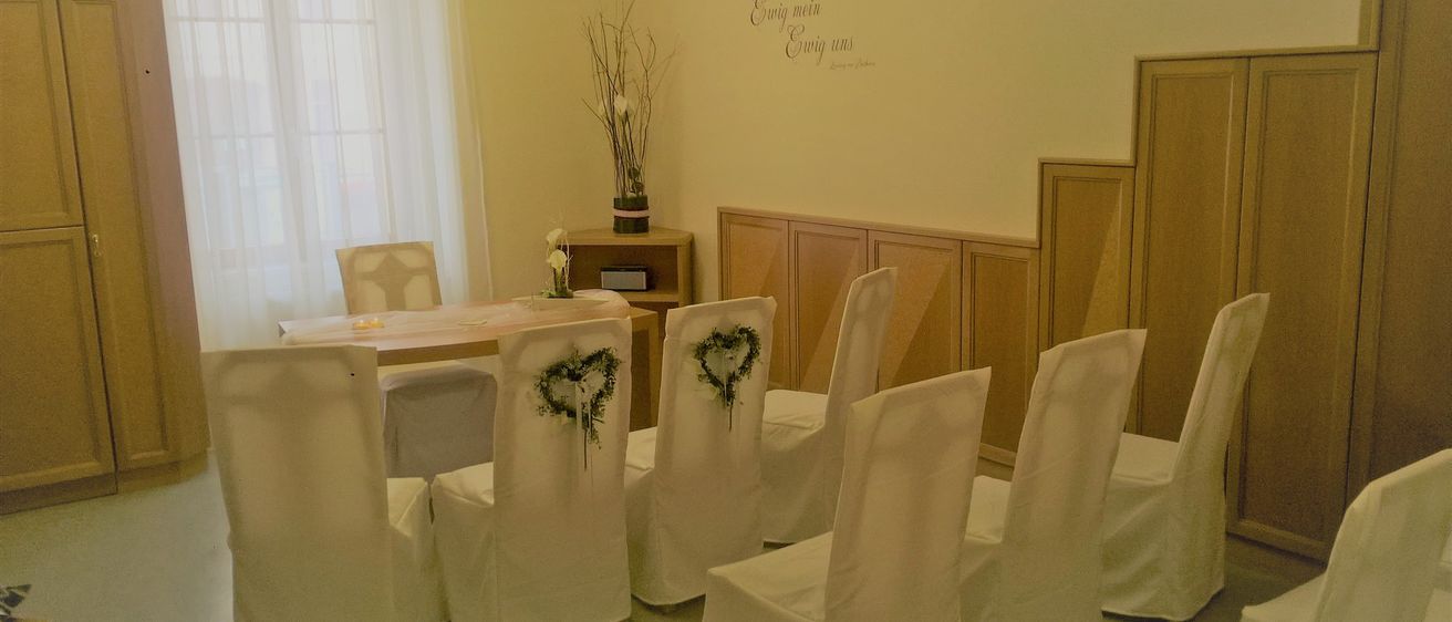 Ein Raum mit weißen Stühlen, einem hölzernen Schreibtisch und einer Wand mit Blumenarrangement und Text. Die Stühle sind in einer Reihe angeordnet, wobei einige dekorative Kränze haben.