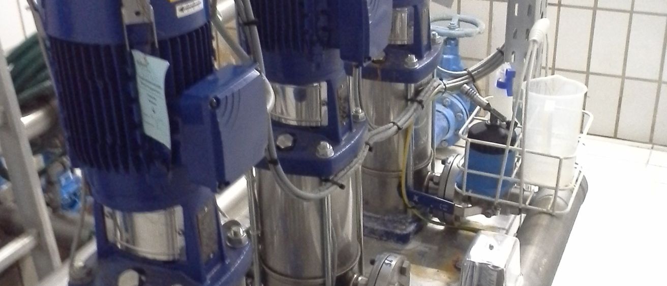 Eine Nahaufnahme von mehreren industriellen Pumpen, alle auf einer Metallplattform mit gefliesten Wänden montiert. Jede Pumpe hat ein blaues und silbernes Gehäuse und ist mit Schläuchen verbunden.