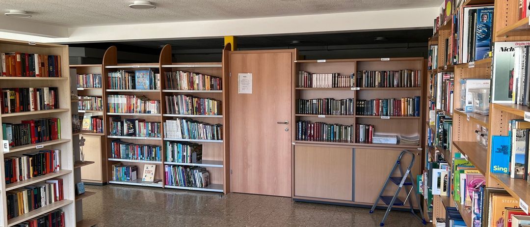Eine leere Bibliothek mit gefliesten Böden und Bücherregalen voller Bücher. Eine Tür befindet sich in der Mitte und eine Leiter ist gegen ein Regal gelehnt.