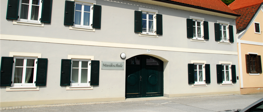 Ein Gebäude mit rotem Dach und grünen Fensterläden, beschriftet mit 'Musikschule', hat mehrere Fenster und eine Tür. Im Hintergrund befinden sich Bäume und ein klarer Himmel.