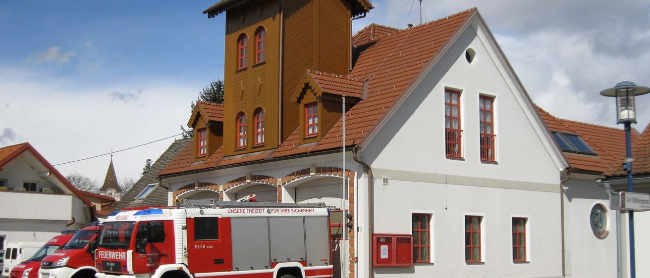 Das Bild zeigt eine Feuerwache mit einem roten und weißen Feuerwehrfahrzeug davor. Das Gebäude hat einen braunen Turm mit roten Fenstern und einem roten Dach. Das Schild zeigt 'Feuerwehr' und 'Neudau', was auf den Standort hinweist. Unten links ist ein Logo zu sehen.