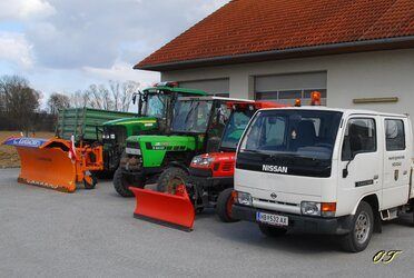 Ein weißer Nissan-Pick-up-Truck ist vor einer Garage mit einem Schneepflug geparkt. Daneben stehen ein roter Traktor, ein grüner Traktor mit Anhänger und ein gelber Traktor.