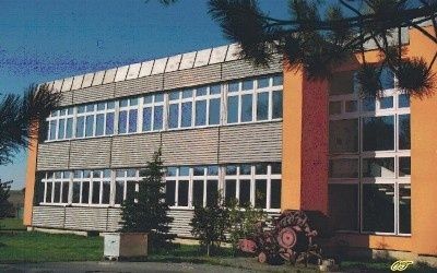 Ein Bürogebäude mit orangefarbenen Wänden und großen Fenstern ist von Pflanzen und Bäumen umgeben. Ein Maschinenteil steht vor dem Gebäude.