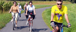 Fünf Menschen fahren auf einem Weg, der von einem Maisfeld umgeben ist. Der Weg wird von Bäumen und Grün beschattet.