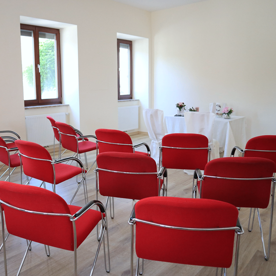 Ein Raum mit weißen Wänden und einem Holzboden. Es gibt rote Stühle, die in Reihen angeordnet sind. Ein Tisch mit einer weißen Tischdecke und Blumen steht im Hintergrund. Es gibt zwei Fenster an der Wand.