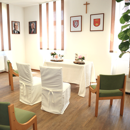 Ein Raum mit einem in weißes Tuch gedeckten Tisch, Blumenarrangements und Kerzen. Vier Stühle umgeben den Tisch, und drei gerahmte Porträts sind an der Wand angebracht.