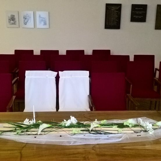 Ein Raum mit purpurroten Stühlen, die in Reihen angeordnet sind, zwei weiße Stühle mit weißen Bezügen und einer Kerze davor, eine weiße Blumendekoration auf einem Holztisch und drei schwarze Plaketten an der Wand.